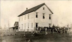 Photograph of Seattle's Belltown School ca. 1881. It is a tall, two-story building the a steep peaked room and a small window in the attic. School children are gathered in front if the building and also looking out from its front windows. Behind the school building on the left there appears to be a house, partially visible. 